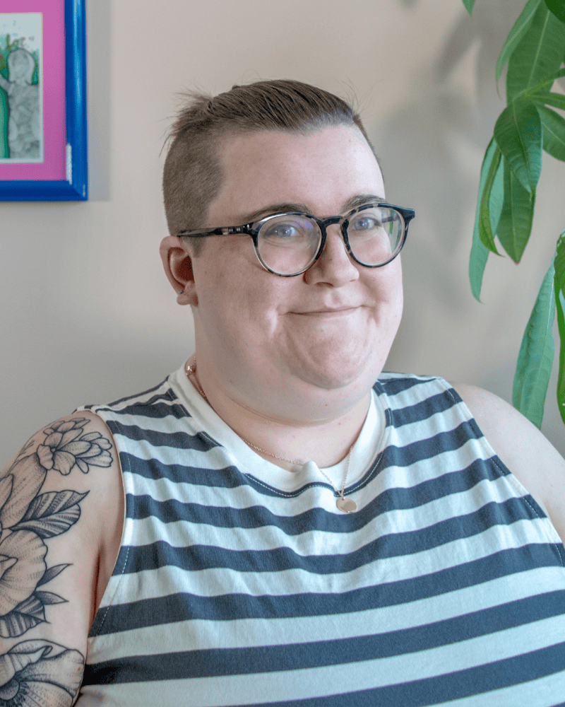 Portrait of a person with short hair and glasses wearing a sleeveless striped shirt, seated indoors near a houseplant with a framed artwork in the background.